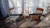 Three children's antique rocking chairs grouped outside on concrete in front of closed garage door. One white painted with thin spindles and decorative motif, one reddish wood with faux covered seat, and one natural wood finish.