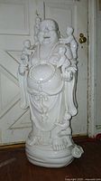 Front view of large white porcelain Buddha statue with five children, smiling face, bead necklace, full robe, standing on rounded base with some damage.