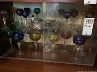 Two glass cabinet shelves displaying coloured cut-to-clear crystal glasses alongside figurines and candle holder
