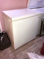 Image showing angled side and top view of white Kenmore Coldspot chest freezer placed on wood floor against pink wall, with black bag nearby.