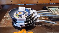 Four stacked frying pans on a wooden surface with visible product labels showing diamond non-stick coating and size details.