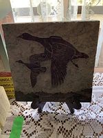 Front view of the square stone plaque showing carved flying geese design on textured stone surface with stand visible behind.