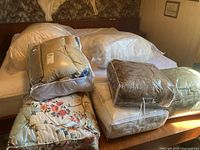 Seven duvets and comforters stacked on a bed, all packaged in plastic bags. Variety of colors and patterns visible including floral and solid light colors.