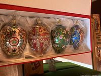 Four decorative Fabergé style ornaments shown in lined red box, individually wrapped in plastic