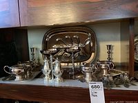 Photo of the silver plate hostess and serving ware arranged on shelf. Includes tray, meat tray with compartments, sugar bowls, creamers, salt and pepper shakers, candlestick holders, small trays and divided serving dish with handle.