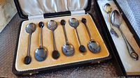 Six sterling silver coffee spoons with black handles displayed in a lined case, together with one sterling silver collector spoon and one additional collector spoon with decorative handle and worn markings.