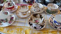 Full view of multiple floral and gold trimmed teacups and saucers on tablecloth