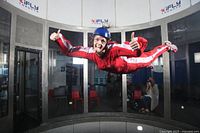 Person in red flight suit and helmet floating inside a vertical wind tunnel giving thumbs up while instructor watches.