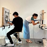 Two people using tufting guns to make rugs on fabric secured by wooden frames.