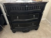 Close-up photo of one of the dark wooden nightstands showing the dark marble top, ornate carved drawer fronts with black inlaid design, and metal ring handles.