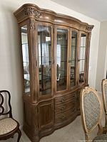 Front-angle view of light wood china cabinet showing curved top, glass doors revealing glass shelves and reflective interior, three central drawers with brass handles, and two lower wooden cabinets with carved details.