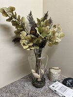 Glass vase with artificial flowers, stems including green leaves and brown feathery foliage, sitting in decorative metal base on a table.