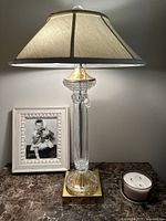 Full lamp with beige fabric shade, clear crystal column, gold square base on marble table with photo frame and candle.
