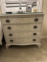 Front view of one white wooden nightstand showing floral pattern on drawer fronts, mirror top, and antique bronze oval drawer pulls