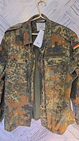 Front view of camouflage military jacket showing button and zipper closure, chest pockets, and collar.
