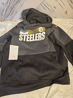 Front view of black Nike hoodie with Pittsburgh Steelers NFL logo including text 'Steelers' and small NFL patch, size tag visible as 6.