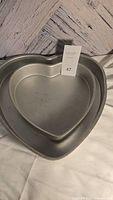 Two nested metal heart-shaped cake molds, the larger approximately 14 inches and the smaller approximately 10 inches, silver color, showing minor wear.