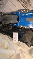 Partial view of blue and gray U.S. Divers fins and black mesh backpack laid on a white surface.
