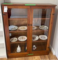 Front view of wooden bookcase cabinet with glass doors, containing decorative plates and vessels on shelves.