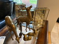 Overview of four brass deer figurines arranged on a glass surface with two glass and metal display cabinets in the background.