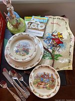 Full set photo showing dinnerware plates and bowls, flatware, apron, Peter Rabbit figure, and booklets on table.