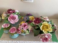 Overview photo showing multiple ceramic floral figurines arranged on a tabletop, featuring roses and carnations in various pastel colors on light blue bases.