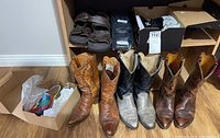 Four pairs of cowboy boots in front, two pairs of brown sandals, pair of black socks, and pair of black sneakers in boxes on shelf behind them.