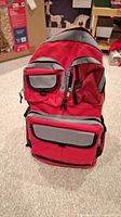 Front view of red and gray emergency preparedness backpack standing upright on carpeted floor.