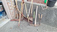Wide view of gardening tools arranged against a brick wall and concrete ground, showing various rakes, shovels, and a pruning saw with visible weathering.