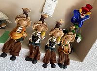 Photo showing four scarecrow shelf-sitters dressed in brown cloth pants with straw hats and smiling faces, arranged sitting on a ledge, and the blue Murano-style glass clown figurine behind them.