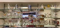 Three glass shelves displaying a variety of clear beer mugs and pint glasses with different brands and designs.