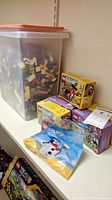 Photo showing a large plastic bin filled with assorted loose Lego bricks of various colors and shapes, along with several boxed Lego sets on a shelf.