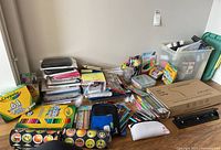 Photo showing a variety of office supplies including notebooks, pencil cases, colored pencils, Crayola crayons and markers, plastic storage boxes, and an LED desk lamp.