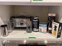 Photo showing all items on a kitchen shelf: DeLonghi slow cooker roaster, NutriBullet blender with cups, Cuisinart burger patty maker box, iSi whipping cream dispenser box, and oil spray bottle box.