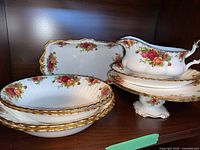Overview of Old Country Roses serving ware including bowls, platter, gravy boat with underplate, and footed compote displayed on shelf