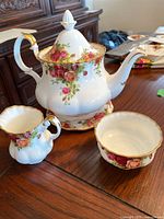 Teapot, creamer, and sugar bowl with floral rose pattern and gold trim on a wooden table.