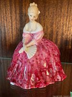 Full front view of the Royal Doulton Victoria figurine on a dark wood shelf showing detailed pink dress and fan.