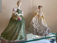 Two Royal Doulton porcelain figurines, Natalie in green dress holding basket and Clarissa in white-yellow dress, standing side by side on a glass shelf.