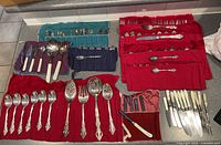 Wide view of multiple sets of silverplated flatware arranged on colored cloth rolls. Shows a variety of spoons, forks, serving spoons, and knives with mother-of-pearl handles.