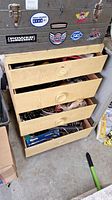 Four-drawer wooden organizer with multiple stickers on metal toolbox above. Drawers partially open showing tools and hardware inside.