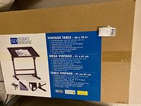 Box showing front and side images of the Studio Designs vintage table including detailed features and angled adjustable surface.