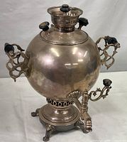 Full view of the antique samovar showing its spherical spout, ornate black-handled side handles, decorative feet, and overall reflective brass body with silver plating.