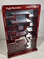 Front view of the wall-mounted curio cabinet showing the reddish-brown stain, glass pane door, tiered shelves, and shell inlay detail on frame edges.