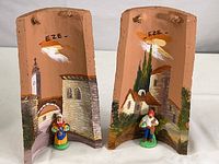 Pair of terra cotta wall hangings with hand painted Eze village scenes and small figurines at base. Both pieces marked 'EZE' at top center. 10 inches tall with 3-inch figurines. Light signs of use.