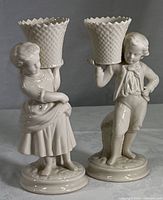 Front view of both porcelain figurines, boy and girl each carrying a basket on their shoulder, standing on circular bases.