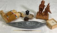 Photo showing full lot including carved stone canoe with paddle, leather moccasins, miniature baskets, metal figurines, small vase, carved seed, wooden box, and inscribed rice grains on white tablecloth.