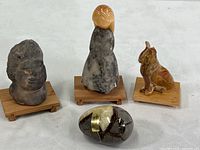 Group photo of four stone figurines and decor items on white surface—primitive carved head, girl figurine, animal figurine, and polished egg.