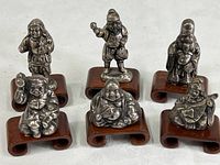 Set of six miniature vintage cast metal Chinese figurines each on individual carved wood platform, arranged in two rows on white cloth background.