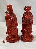 Pair of red cinnabar style Chinese figurines, male on left with broken sword handle piece in front, female on right holding a basket, both standing on detailed bases.