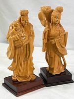 Two hand-carved Chinese wood figurines, a man holding a baby and a woman, shown together on wooden bases.